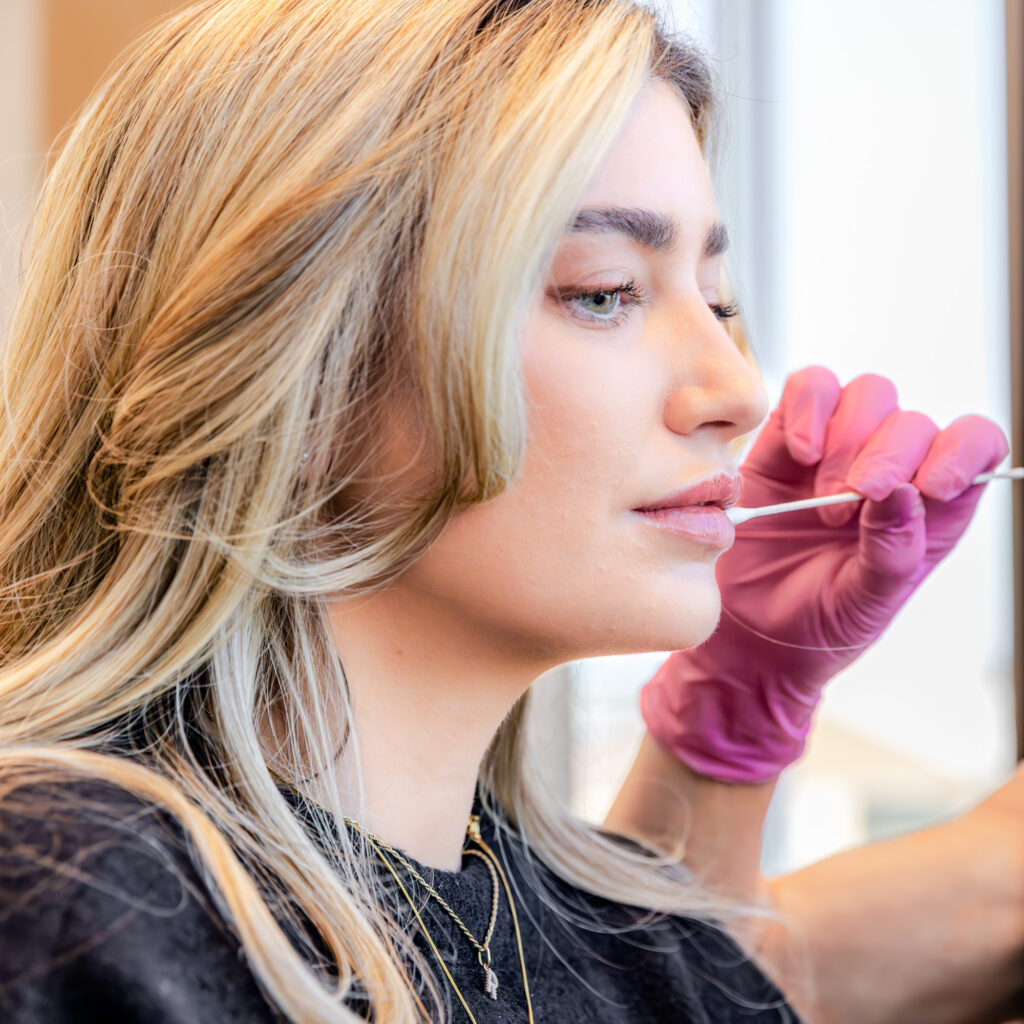 Patient looking straight ahead as her esthetician holds a q-tip up to her lips and assess whether the patient would benefit from a lip flip or lip filler in Plano.