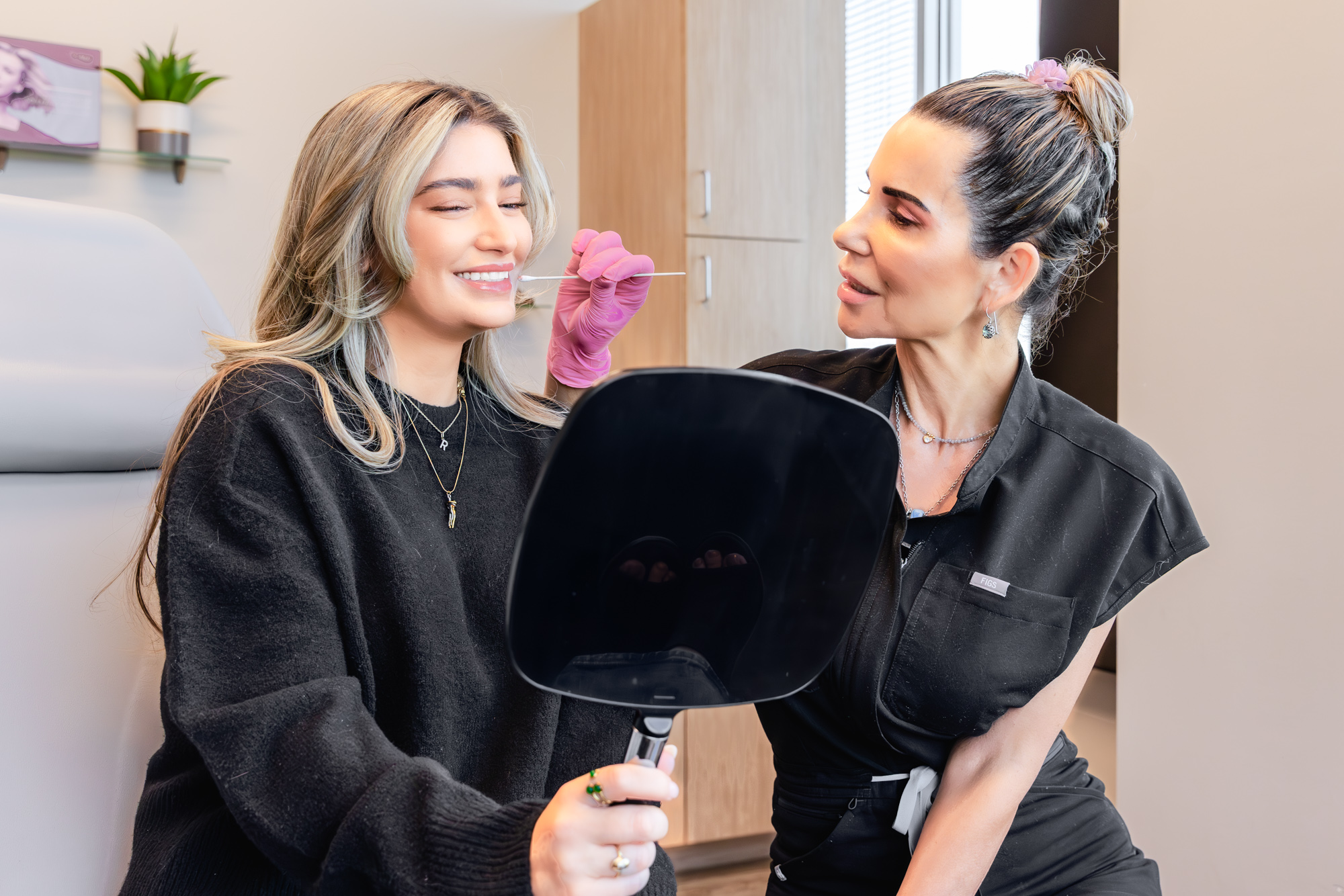 Patient smiles and looks in a handheld mirror while plastic surgeon holding cotton swab holds it near her face, assessing the best placement on the patient's jawline for chin filler near Frisco