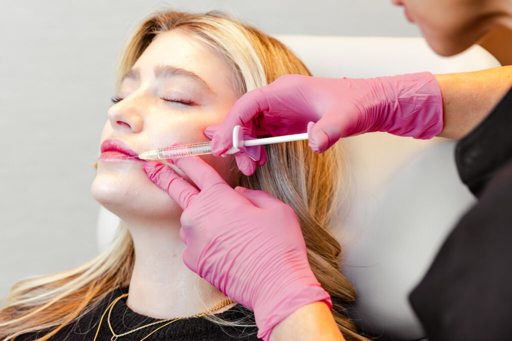 Female patient leaning back as an esthetician wearing gloves injects her lips with dermal filler in Plano.