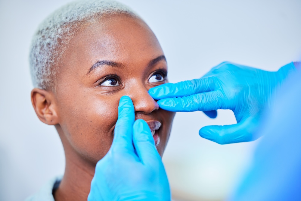 Woman smiles and looks up as plastic surgeon places their finger on both sides of her nose to determine the best treatment plan for her nose job in Plano