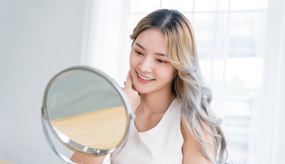 Young woman with glowing skin looking in mirror and admiring her complexion after getting a treatment of Clear and Brilliant laser near Prosper.