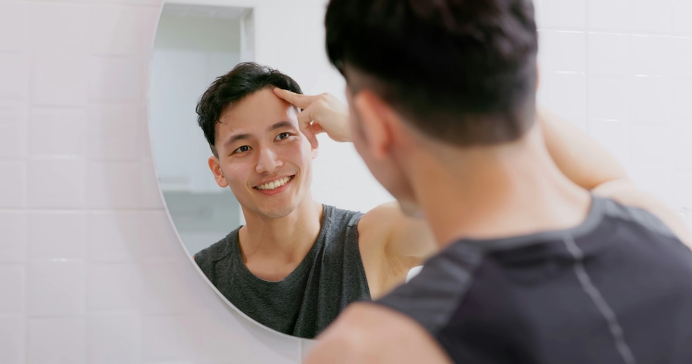 Young man looking at himself confidently in the mirror. He is noticing renewed hair growth after starting treatment for hair restoration for men in Plano.